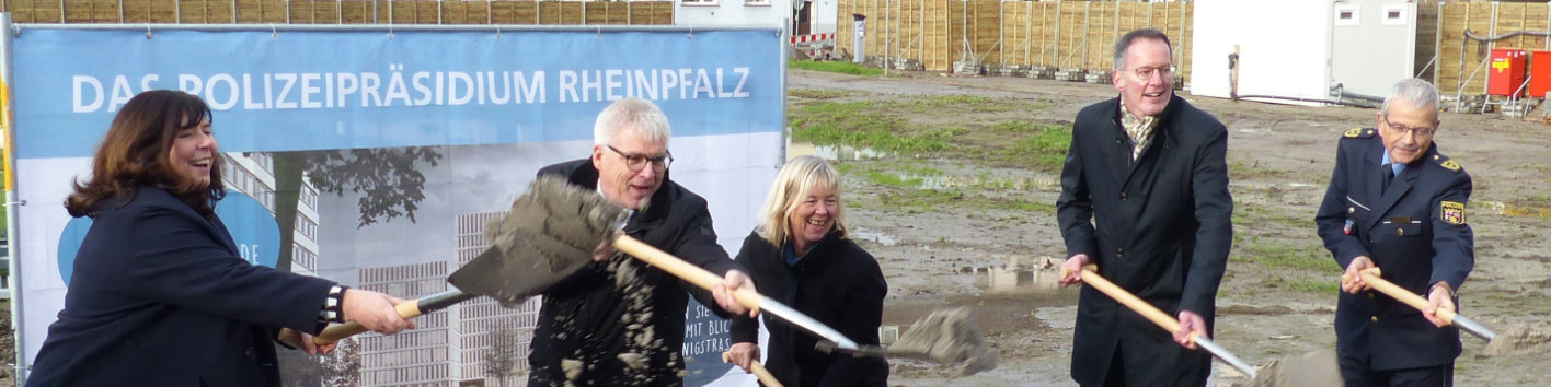 Fünf Menschen mit Spaten in der Hand stehen vor einer Plakatwand auf einer Baustelle.