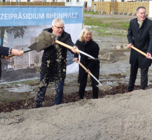 Fünf Menschen mit Spaten in der Hand stehen vor einer Plakatwand auf einer Baustelle.