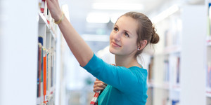 Eine junger Frau, die einen türkis-blauen Pullover trägt, sucht nach einem Buch in einer Bibliothek