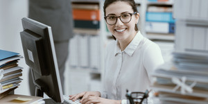 Eine junge, lächelnde Verwaltungskraft sitzt in einem Büro vor einem PC und lacht in die Kamera