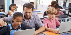 Ein Lehrer im Unterricht erklärt einer Schülern eine Aufgabe am Notebook. Die anderen Schülerinnen und Schüler arbeiten auch mit dem Notebook